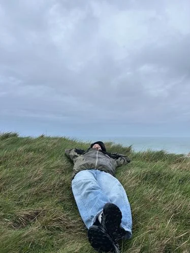 A person lies in tall grass on a cliff, looking out toward a foggy sea.
