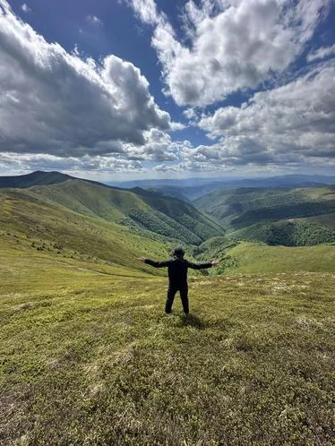 A figure stands with arms outstretched on a green mountain ridge, overlooking a valley.